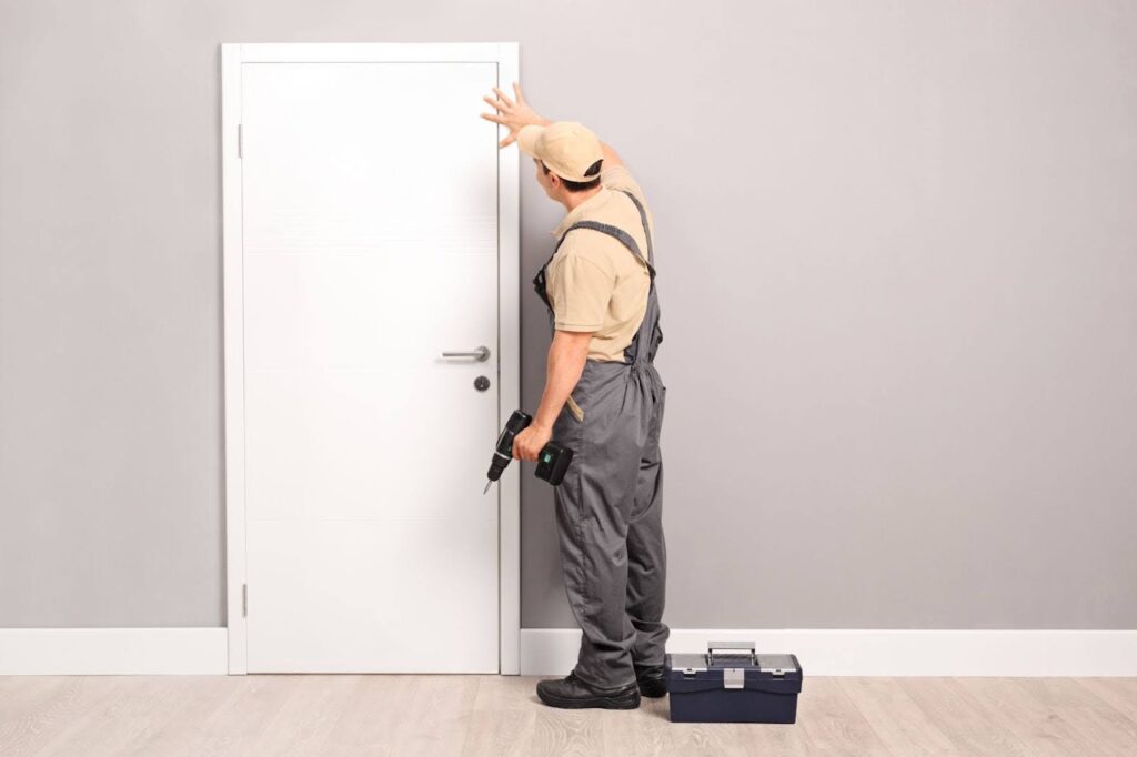 A plain white door being installed by a young handyman holding a drill.